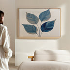 A person in white stands in a minimalist room, admiring the Sapphire Leaves Canvas Art - Horizontal. In front is a light sofa with wooden armrest, perfectly complementing the tranquil, botanical vibe of the blue leaf wall art.