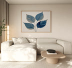 A modern living room features a light beige sectional, low wooden coffee table, potted plant, and the Sapphire Leaves Canvas Art - Horizontal as striking blue wall art above the couch. Natural light brightens the space.