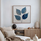 A modern, minimalist living room with a beige sofa, neutral cushions, and a wooden sideboard featuring the Sapphire Leaves Canvas Art - Square in blue and gray tones. Decorative vases and books complete the serene botanical decor.