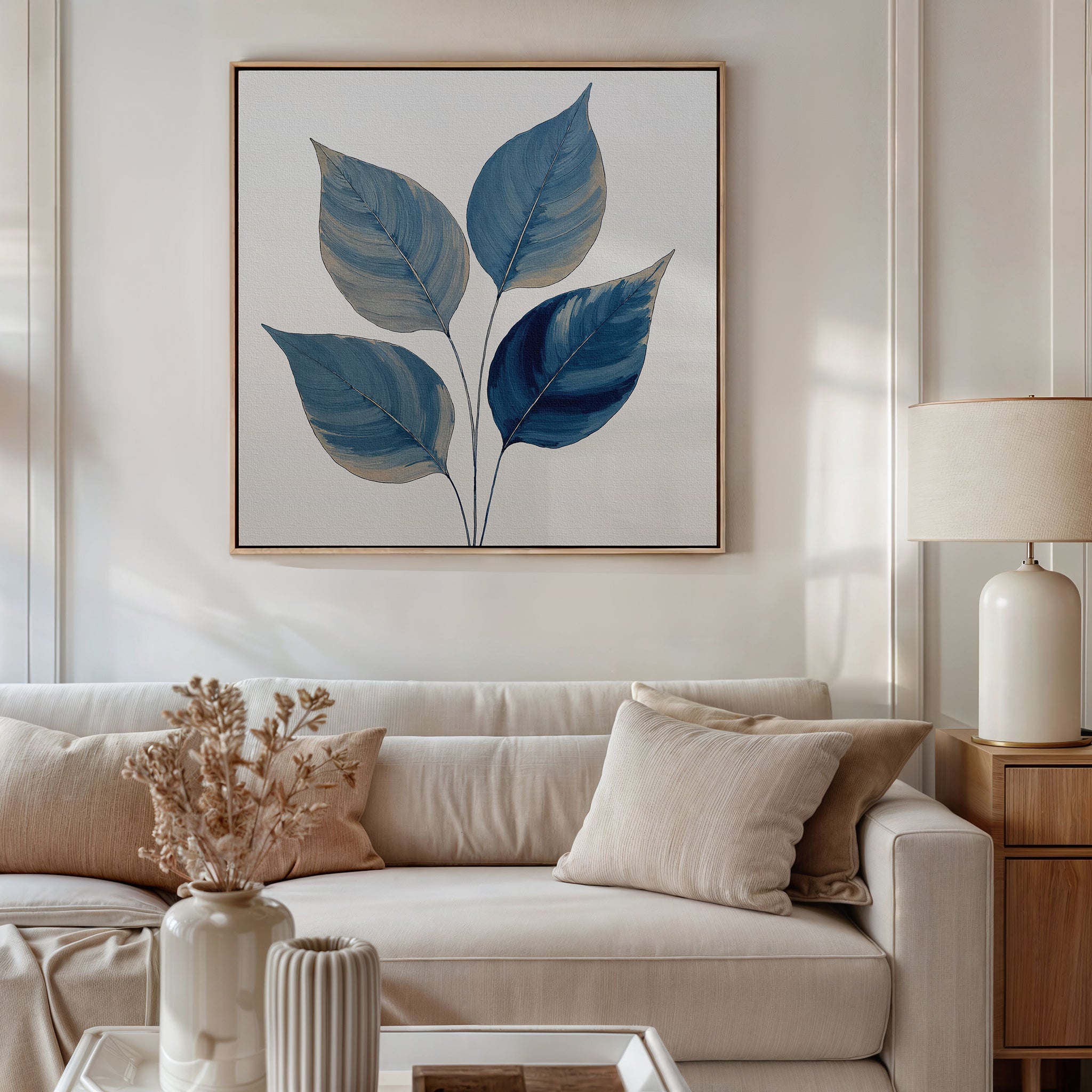 A neutral-toned living room features a beige sofa, cream cushions, and a light wood side table with a white lamp. Above the sofa hangs the Sapphire Leaves Canvas Art - Square, adding botanical charm, while dried flowers decorate the coffee table.