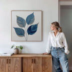 A woman in a white blouse and jeans leans against a wooden cabinet, gazing at the Sapphire Leaves Canvas Art - Square with botanical blue leaves. A potted plant and book sit on the cabinet beneath the artwork.