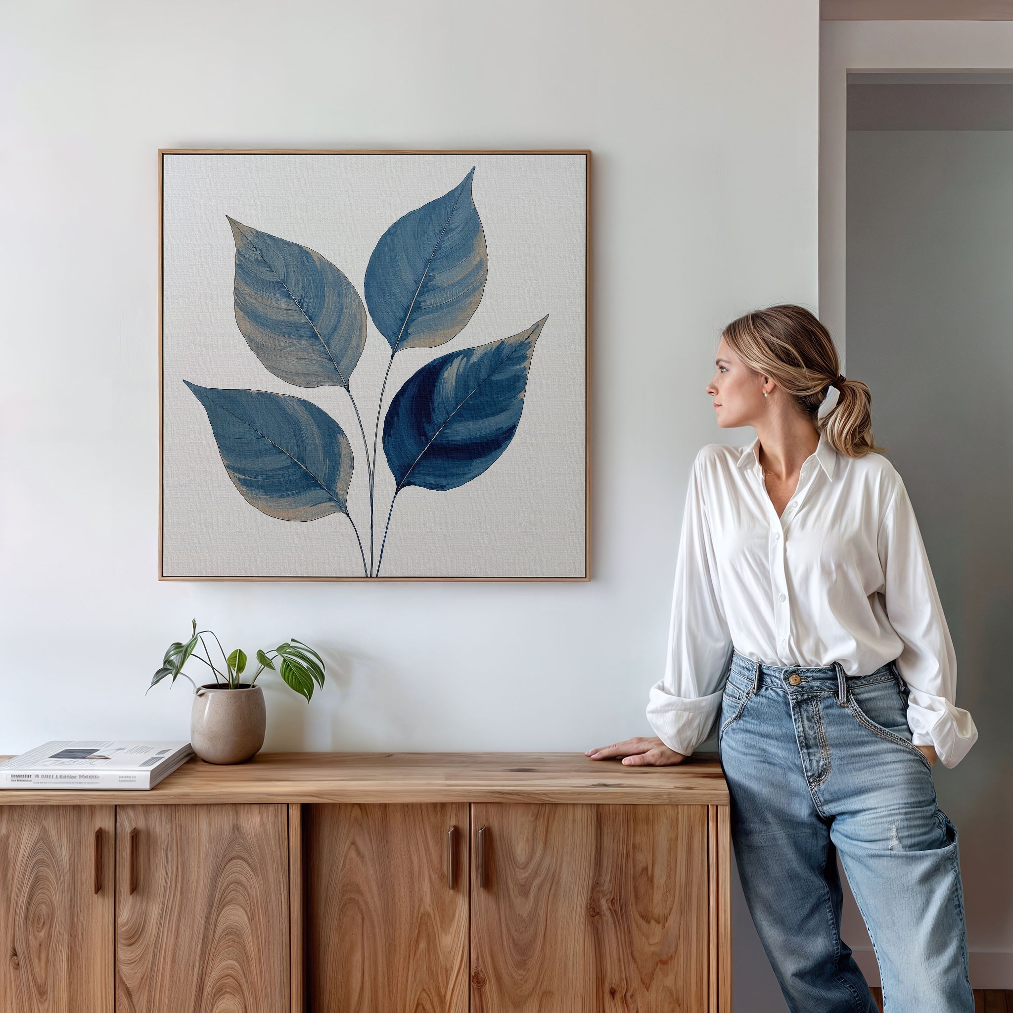 A woman in a white blouse and jeans leans against a wooden cabinet, gazing at the Sapphire Leaves Canvas Art - Square with botanical blue leaves. A potted plant and book sit on the cabinet beneath the artwork.