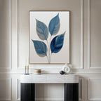 A modern room features calming blue wall art—a Sapphire Leaves Canvas Art - Vertical displayed above a white marble console table, which holds decorative objects like candles, a round sculpture, and a lidded container.