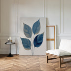 A modern room with white paneled walls, herringbone wood floor, a cushioned chair, round black side table with candles and books, and the calming Sapphire Leaves Canvas Art - Vertical botanical print resting against the wall.