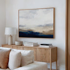 A modern living room features a light wood sideboard with stacked books, a lamp, and a bowl. Above it hangs the Sapphire Tides Canvas Art - Horizontal, a large abstract print in cobalt blue, white, and beige tones. A white sofa is partially visible in front.