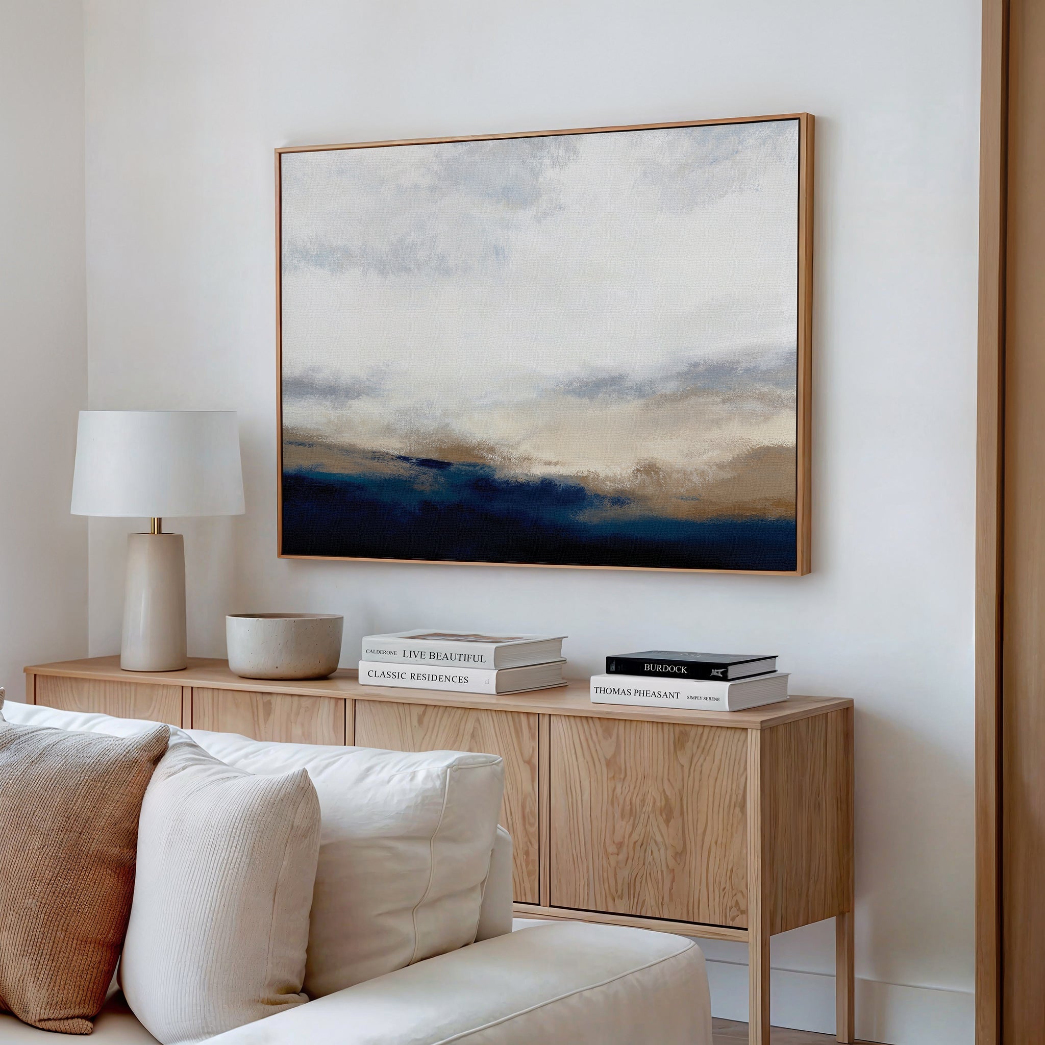 A modern living room features a light wood sideboard with stacked books, a lamp, and a bowl. Above it hangs the Sapphire Tides Canvas Art - Horizontal, a large abstract print in cobalt blue, white, and beige tones. A white sofa is partially visible in front.