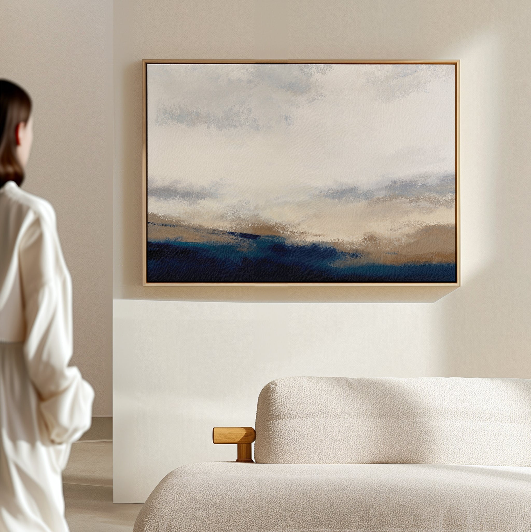 A person in a white shirt stands by a beige sofa, admiring the Sapphire Tides Canvas Art - Horizontal, a framed abstract print with cobalt blue and beige tones that adds modern wall decor to the light-colored room.