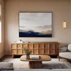 A modern living room with a light sofa, wooden coffee table, and carved sideboard features Sapphire Tides Canvas Art - Horizontal above. Blue, beige, and white details plus a small vase and books finish the decor.