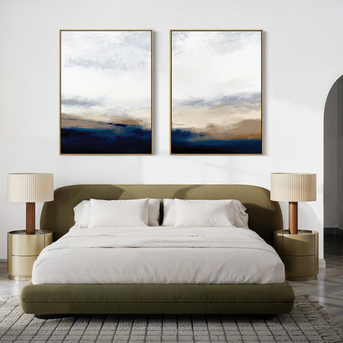 A modern bedroom with an olive green bed, white bedding, gold nightstands, beige lamps, and Sapphire Tides - Set Of 2 abstract landscape prints in subtle cobalt blues hanging above the bed.