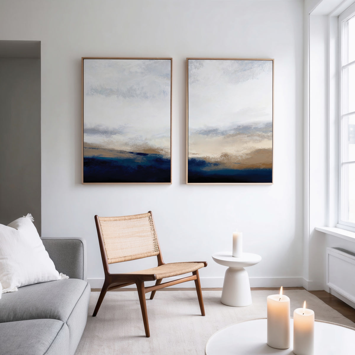 A modern living room showcases a wooden chair, gray sofa with white pillows, and a small white side table. Sapphire Tides - Set Of 2 abstract prints in cobalt blue hang on the wall above three lit candles on a low table.