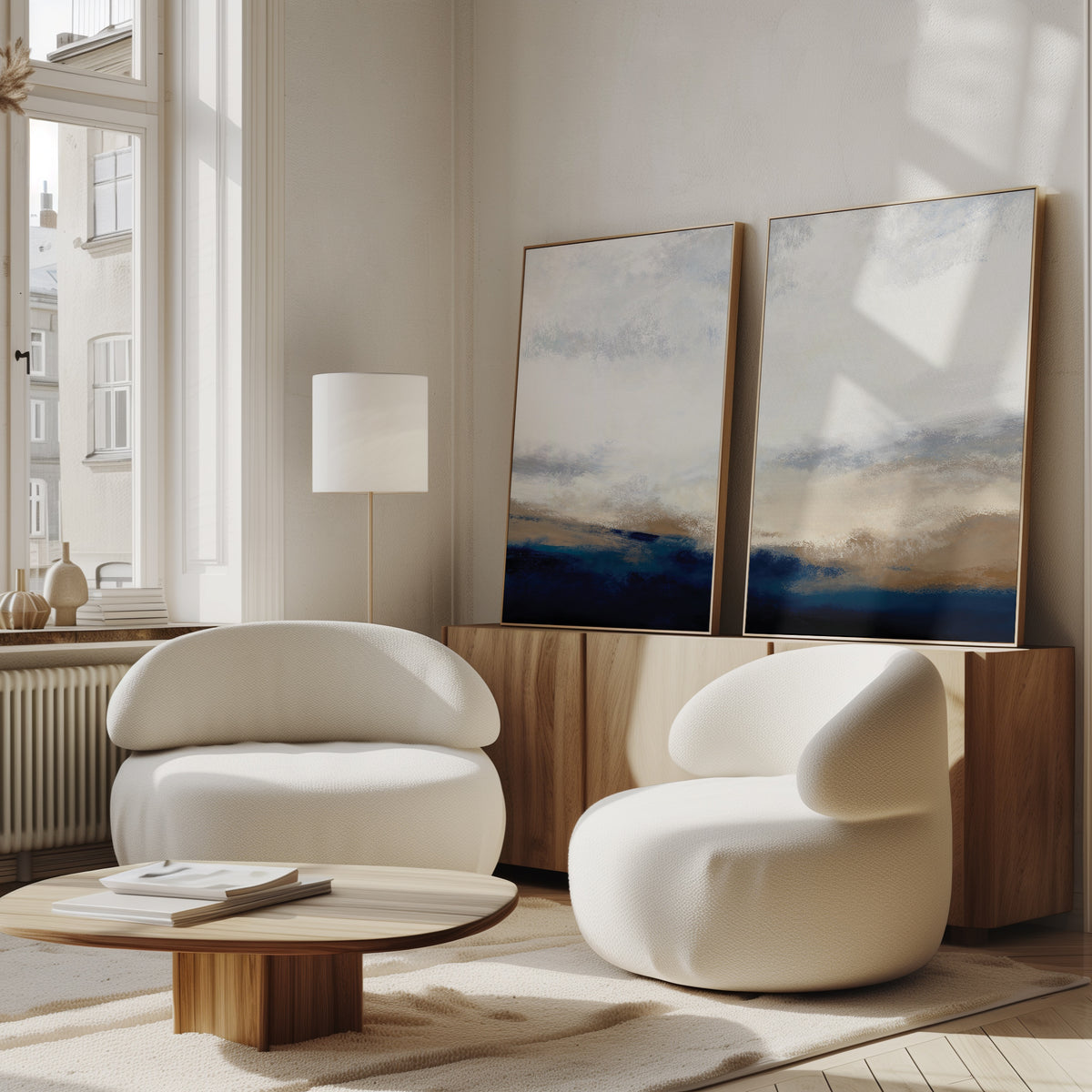Bright, modern living room with two rounded white chairs, a low wooden coffee table, a tall floor lamp, and the Sapphire Tides - Set Of 2 artwork in cobalt blues leaning on a wooden cabinet, sunlight streaming through large windows.