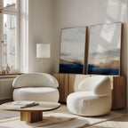 Bright, modern living room with two rounded white chairs, a low wooden coffee table, a tall floor lamp, and the Sapphire Tides - Set Of 2 artwork in cobalt blues leaning on a wooden cabinet, sunlight streaming through large windows.