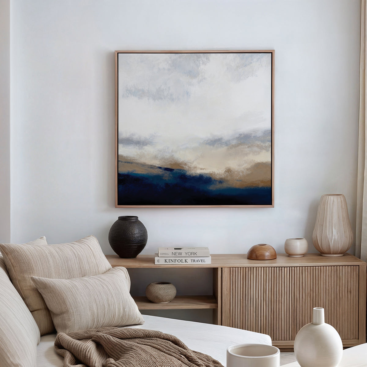 A minimalist living room displays the Sapphire Tides Canvas Art - Square in cobalt blues, brown, and white above a beige sofa with striped pillows and a light wooden sideboard—perfect for modern spaces.
