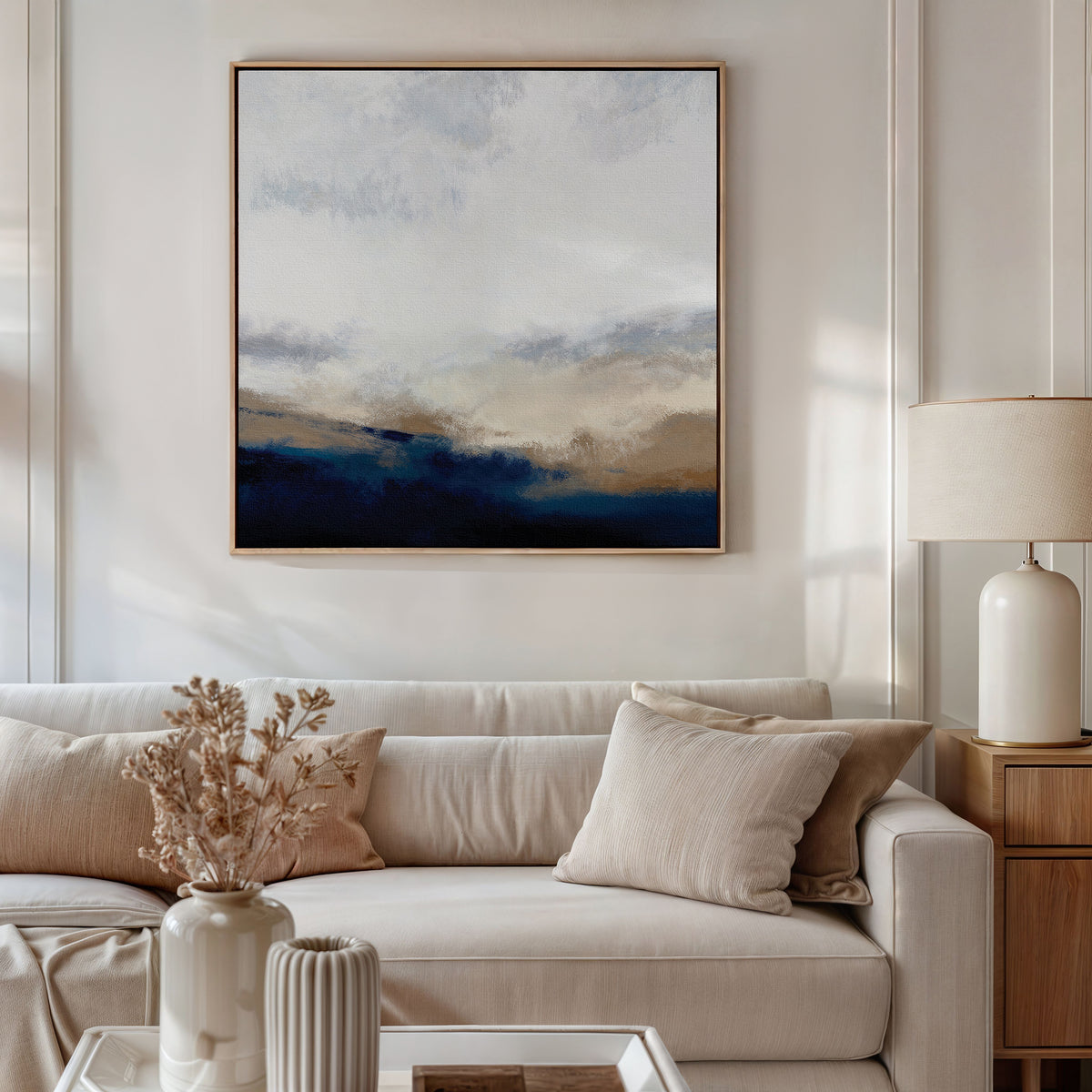 A modern living room with a light beige sofa, cream pillows, a side table with a ceramic lamp, dried flowers in a vase, and the Sapphire Tides Canvas Art - Square featuring cobalt blue, beige, and white tones on the wall.