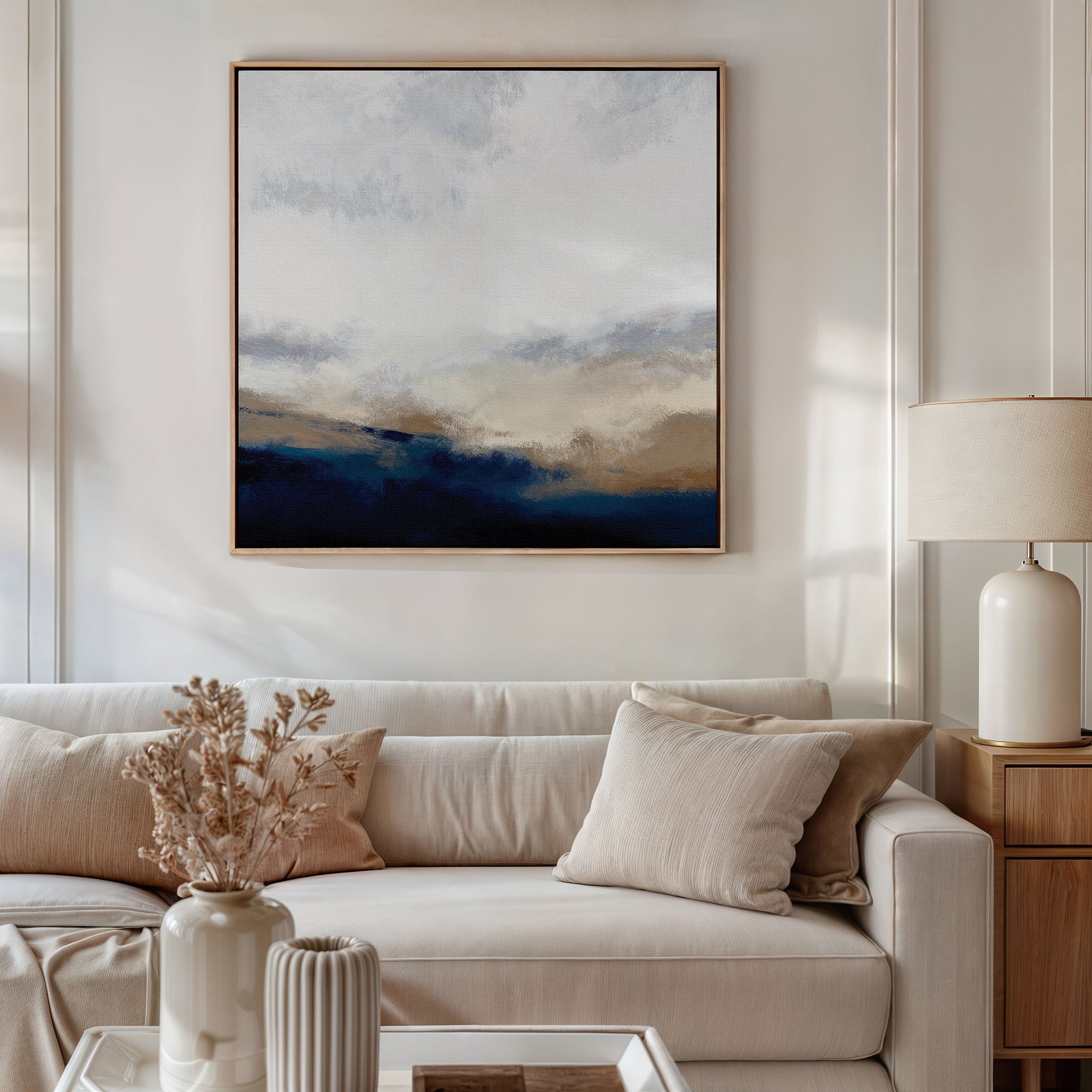 A modern living room with a light beige sofa, cream pillows, a side table with a ceramic lamp, dried flowers in a vase, and the Sapphire Tides Canvas Art - Square featuring cobalt blue, beige, and white tones on the wall.