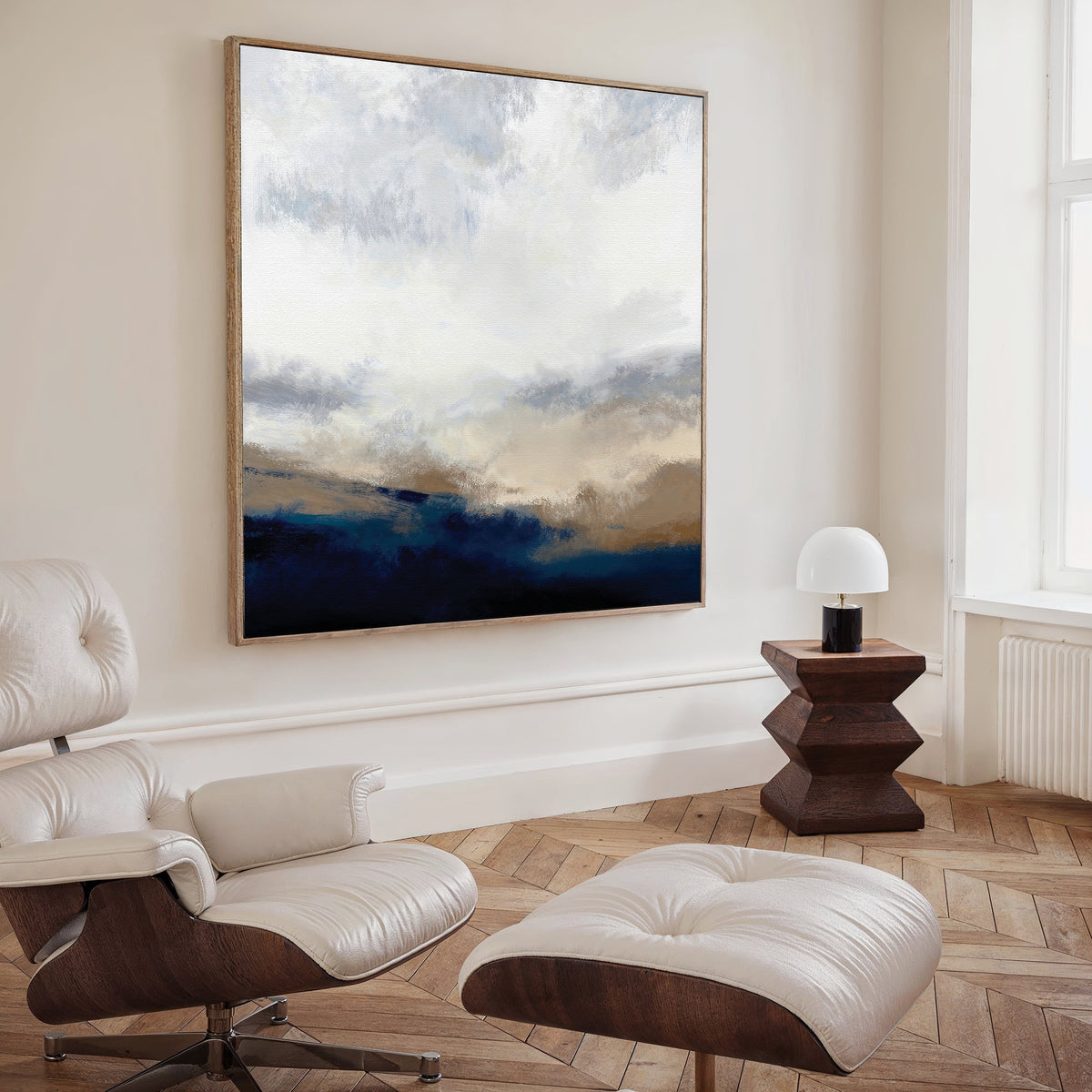 A modern living room featuring a white leather lounge chair and ottoman, wooden side table with a white lamp, and the Sapphire Tides Canvas Art - Square showcasing cobalt blue, beige, and white tones on a cream-colored wall.