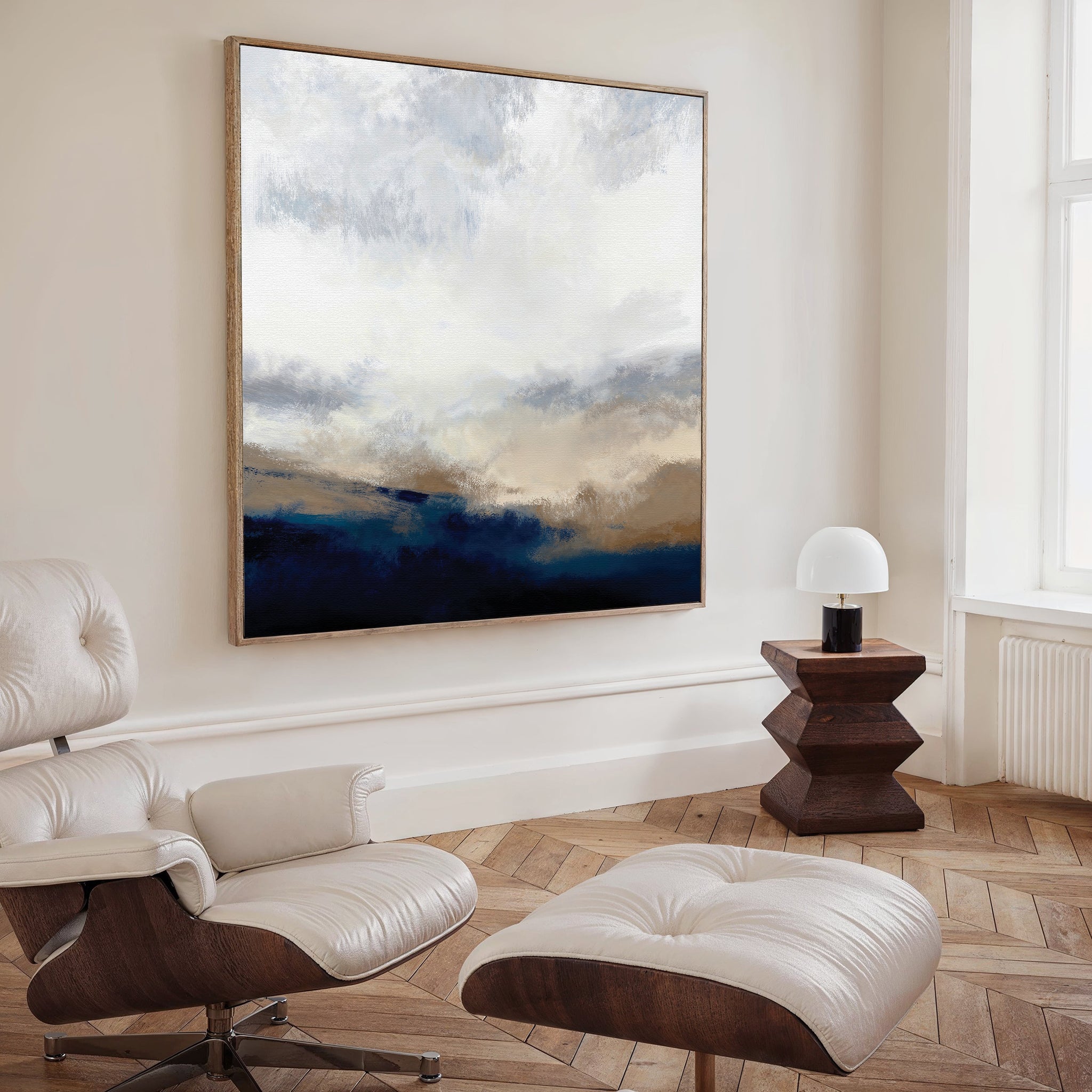 A modern living room featuring a white leather lounge chair and ottoman, wooden side table with a white lamp, and the Sapphire Tides Canvas Art - Square showcasing cobalt blue, beige, and white tones on a cream-colored wall.