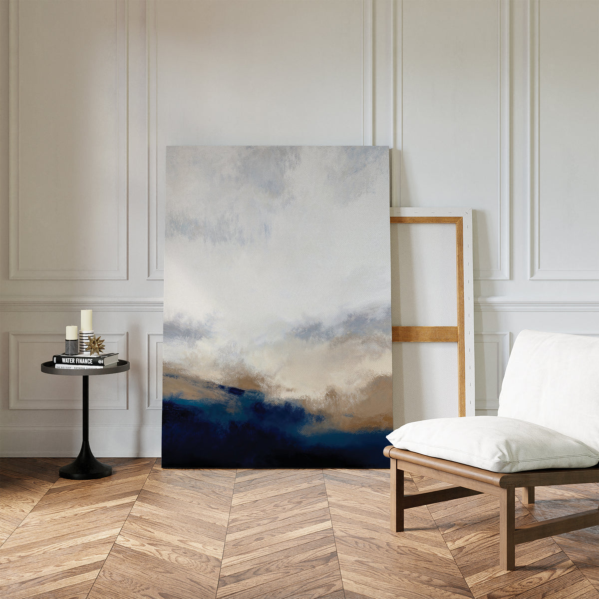 The Sapphire Tides Canvas Art - Vertical in cobalt blue and beige leans against a white paneled wall, ideal for modern living rooms. Nearby are an empty wooden frame, candles, books on a black table, and a cushioned chair on chevron wood floors.