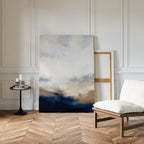 The Sapphire Tides Canvas Art - Vertical in cobalt blue and beige leans against a white paneled wall, ideal for modern living rooms. Nearby are an empty wooden frame, candles, books on a black table, and a cushioned chair on chevron wood floors.