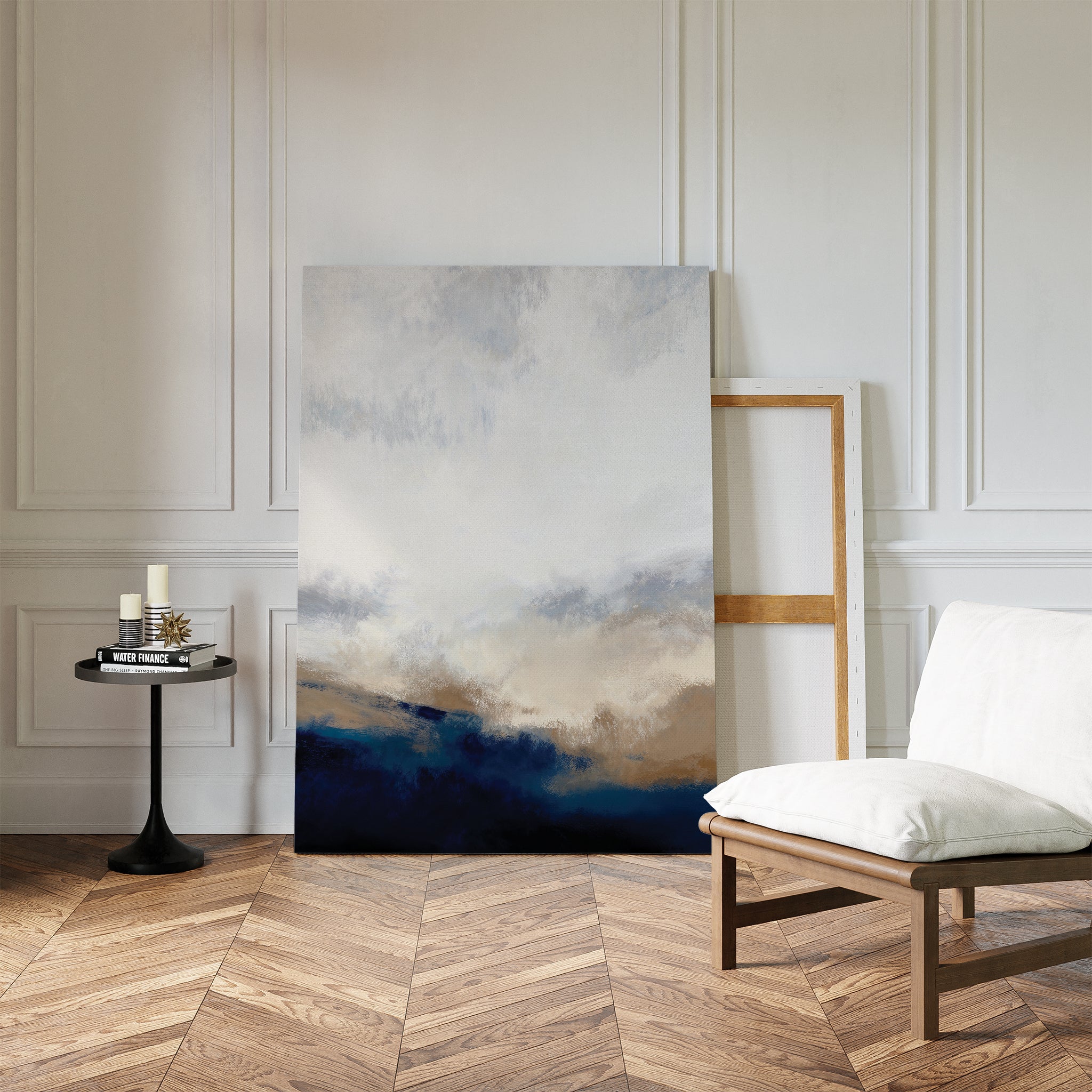 The Sapphire Tides Canvas Art - Vertical in cobalt blue and beige leans against a white paneled wall, ideal for modern living rooms. Nearby are an empty wooden frame, candles, books on a black table, and a cushioned chair on chevron wood floors.