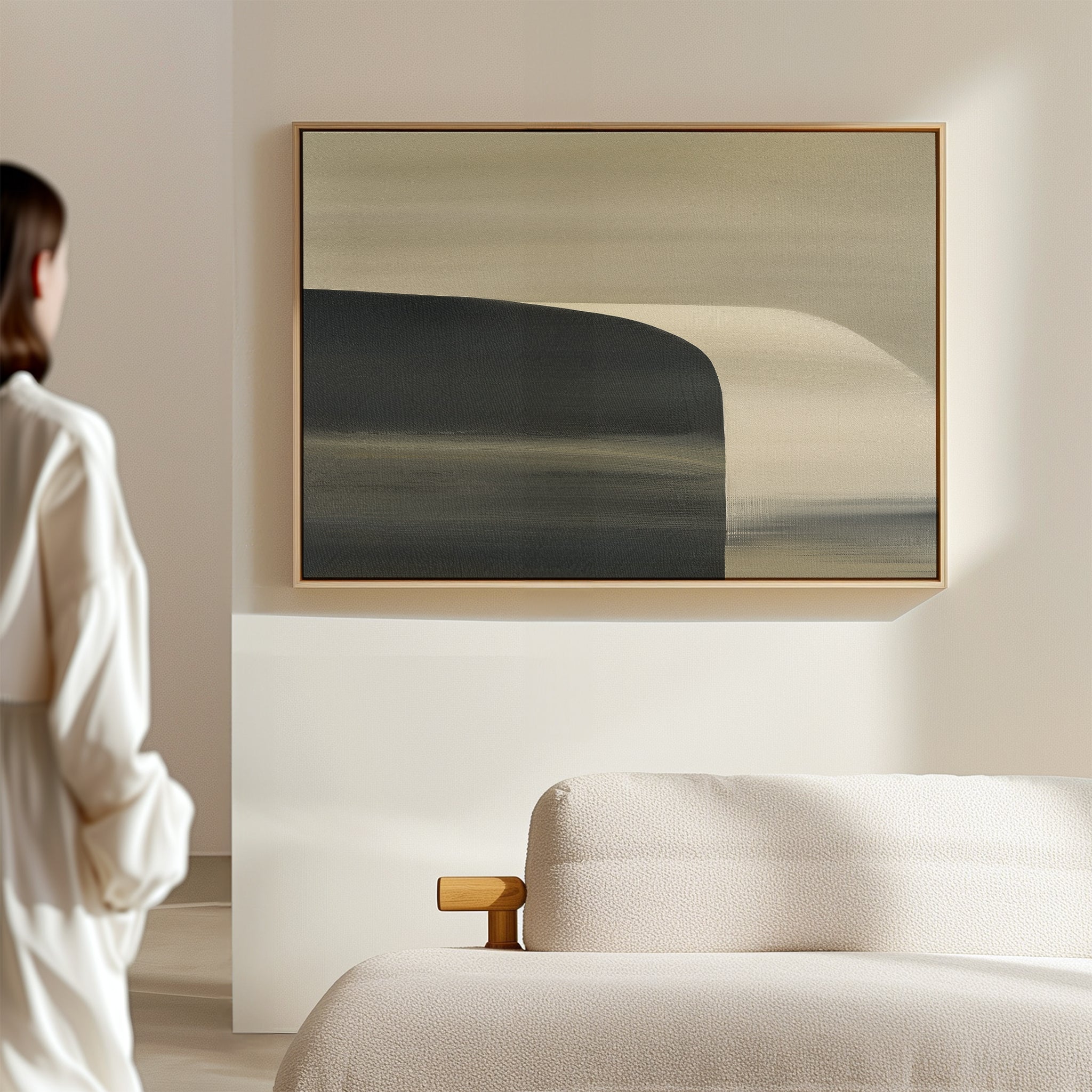 A person in a white outfit admires the Sculptural Forms Canvas Art - Horizontal, featuring soft, muted colors above a beige textured couch in a minimalistic, sunlit living room with modern decor.