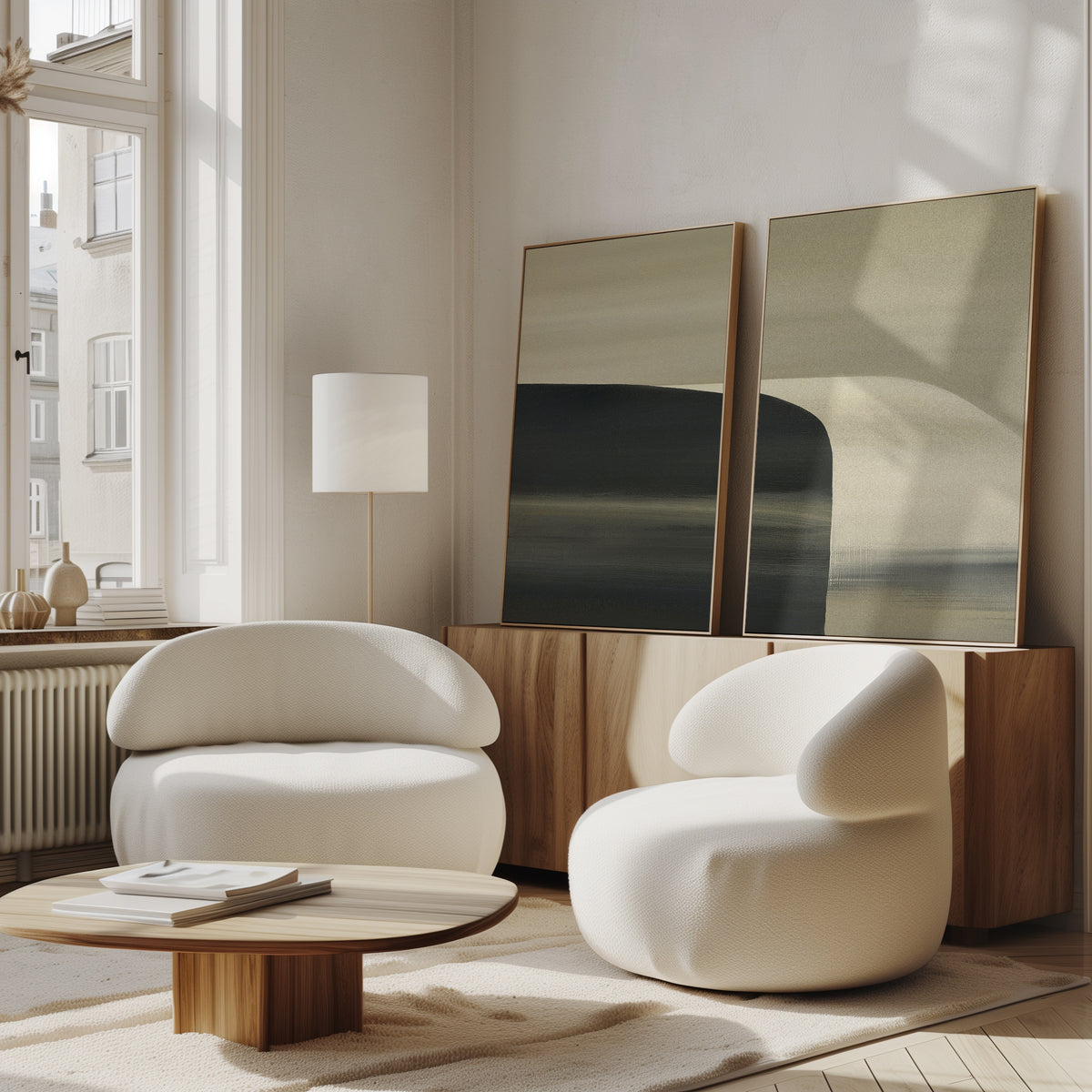 A modern living room features Sculptural Forms - Set Of 2 white armchairs, a low wooden coffee table, and a sideboard with abstract art. Large windows flood the space with natural light, highlighting the minimalist and neutral design.