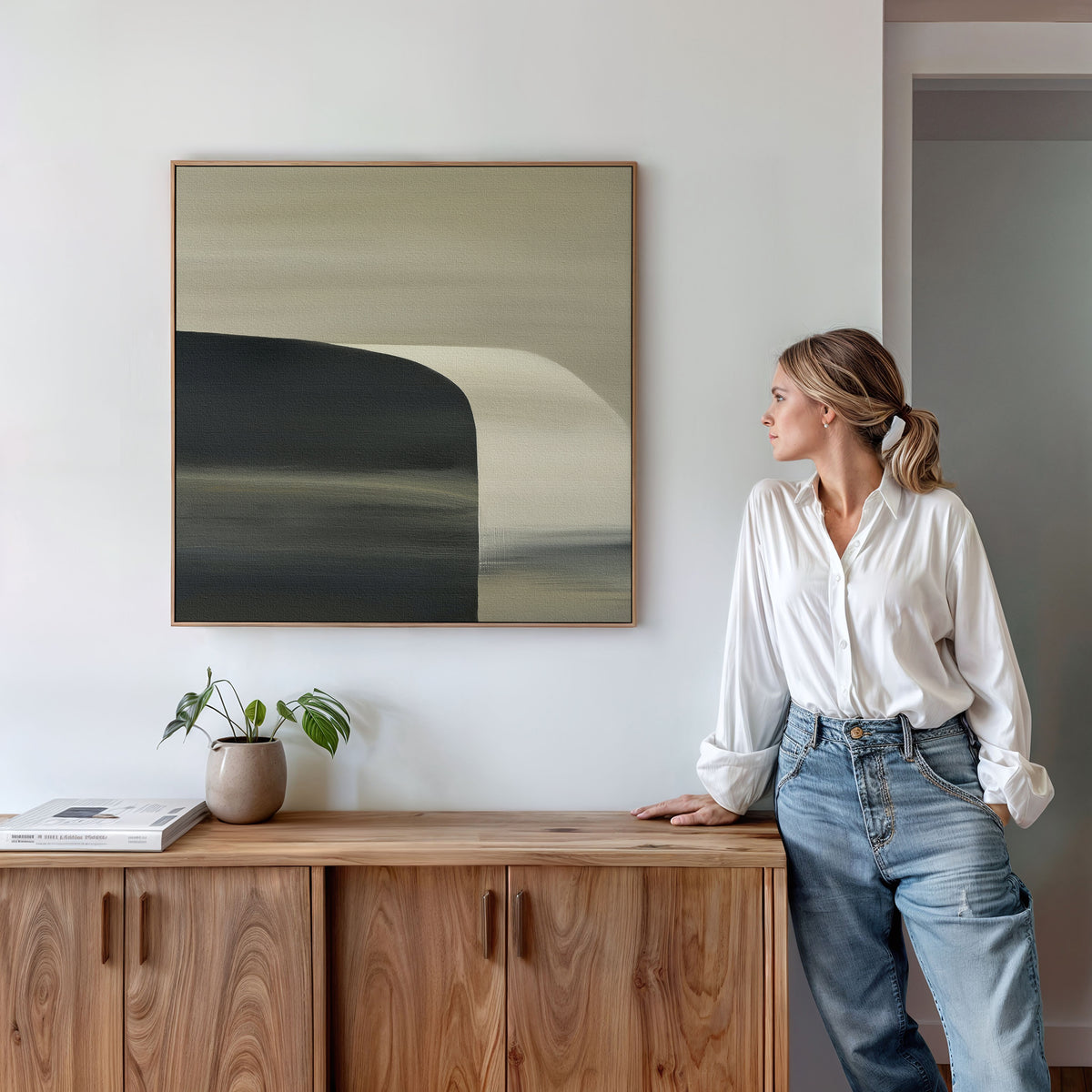 A woman in a white blouse and jeans stands beside a wooden cabinet, admiring the Sculptural Forms Canvas Art - Square. Soft earthy tones, a small plant, and books complete the minimalist decor, creating a tranquil modern living room ambiance.