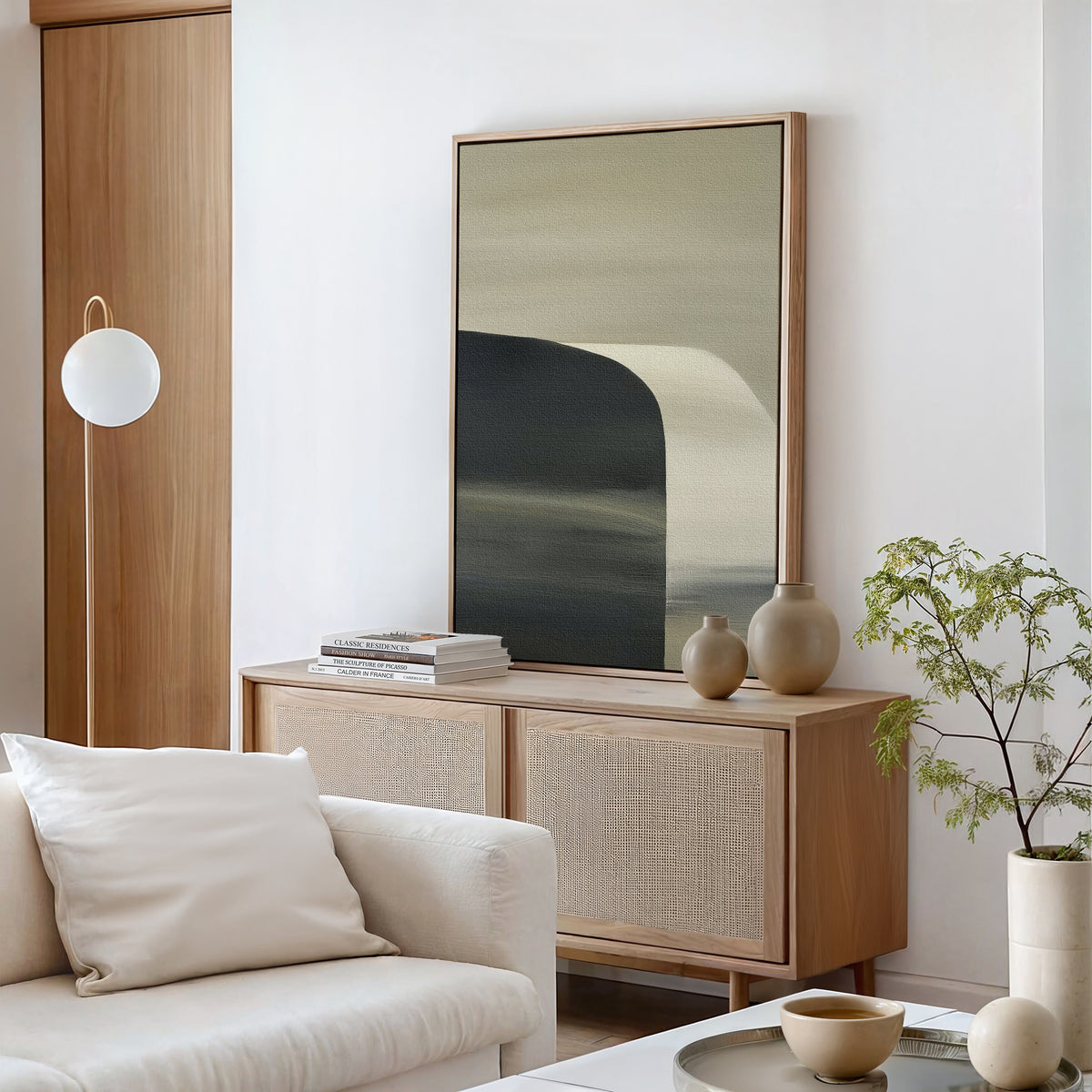 A minimalist living room with light wood furniture, a white sofa, a modern sideboard, ceramic vases, stacked books, a potted plant, and a round hanging light features the Sculptural Forms Canvas Art – Vertical as a striking focal point.