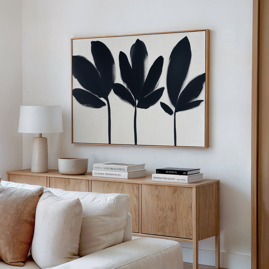 A modern living room features a light wood sideboard with neutral decor, books, and a white table lamp. Above it hangs the Shadow Stems Canvas Art - Horizontal with bold botanical silhouettes. A beige sofa sits in the foreground.