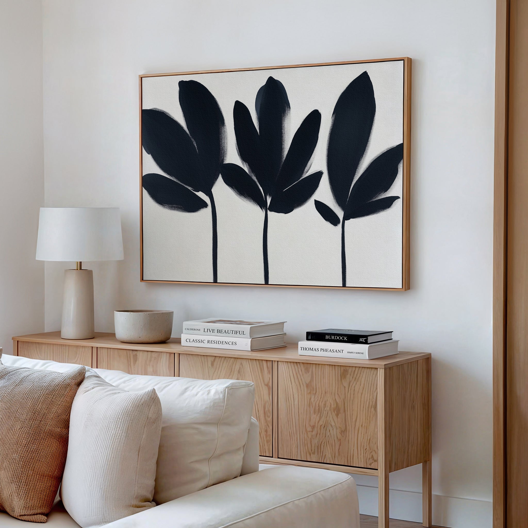 A modern living room features a light wood sideboard with neutral decor, books, and a white table lamp. Above it hangs the Shadow Stems Canvas Art - Horizontal with bold botanical silhouettes. A beige sofa sits in the foreground.