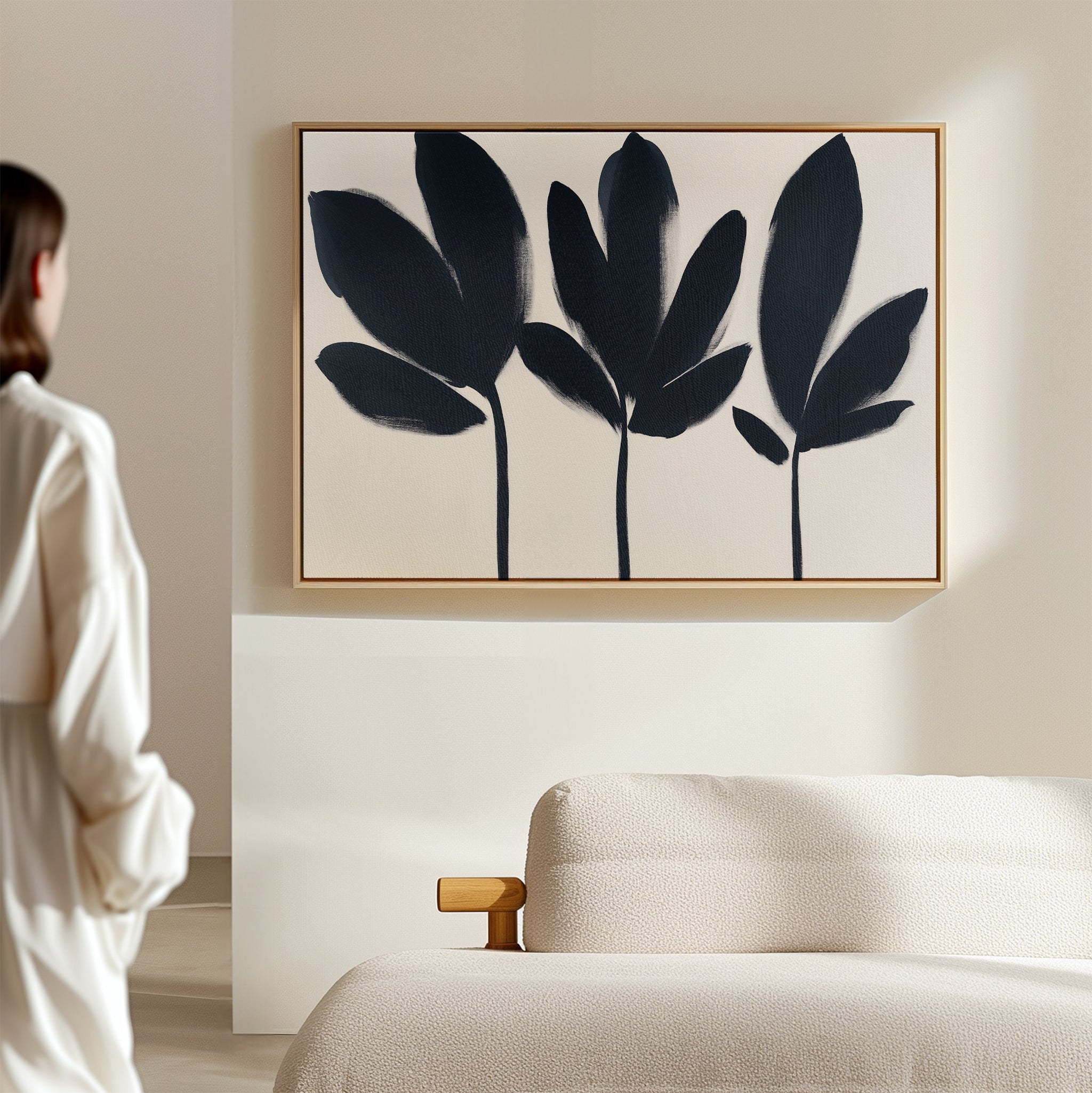 A person in a white shirt admires the Shadow Stems Canvas Art - Horizontal, showcasing minimalist black botanical silhouettes, displayed above a light sofa in a modern living room.