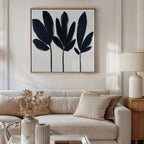 A modern living room with a beige sofa, cream cushions, wooden side table and lamp, vase of dried flowers, and the Shadow Stems Canvas Art - Square with black-and-white botanical silhouettes displayed on the wall.