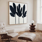 A modern, minimal living room features a white lounge chair with ottoman, wooden side table with a small white lamp, and the Shadow Stems Canvas Art - Square—a large black-and-white botanical print—on the wall.