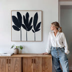 A woman in a white blouse and jeans leans against a wooden cabinet, admiring the Shadow Stems Canvas Art - Square. The minimalist botanical print adds chic style to her modern living room, complemented by a potted plant and a book for serenity.