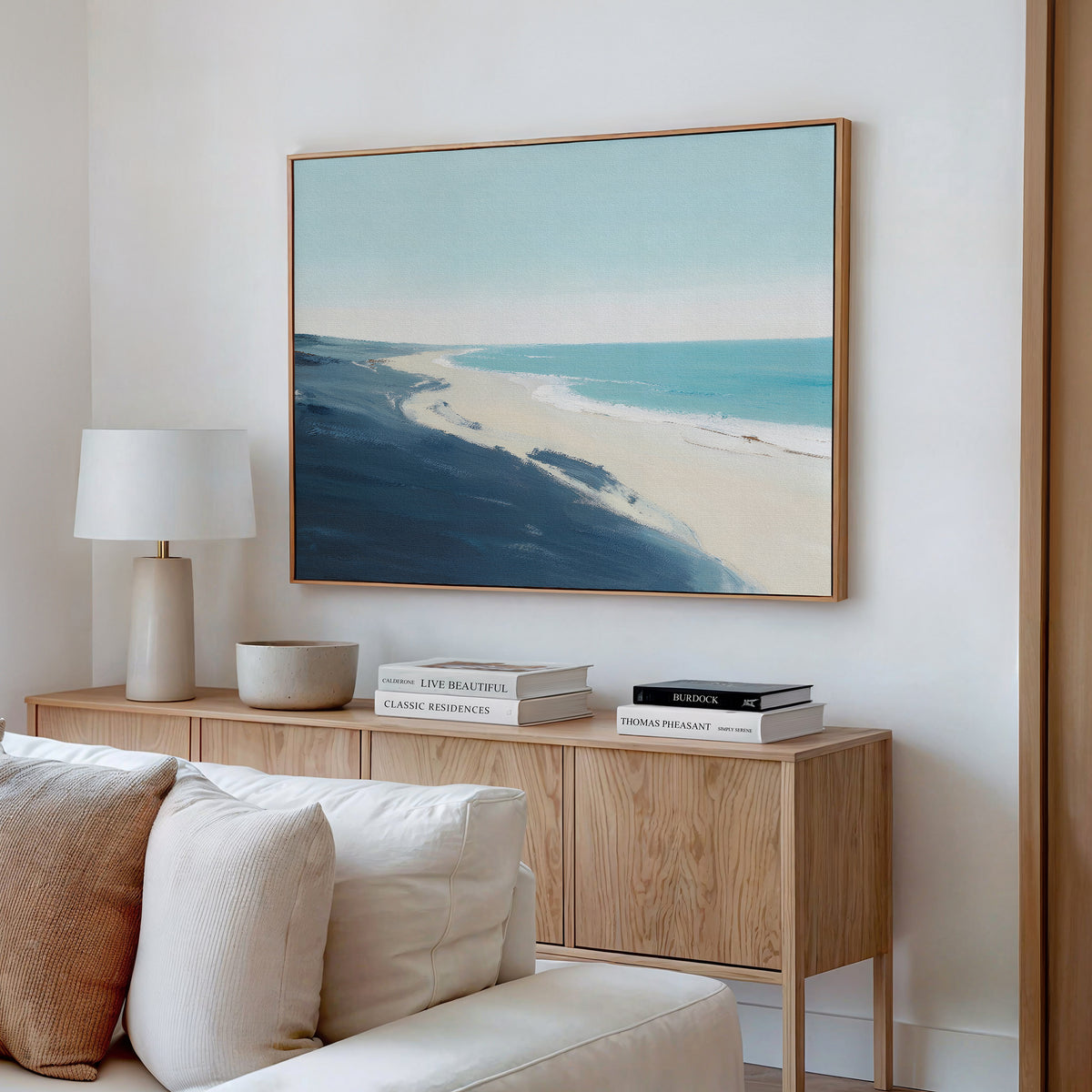 A modern living room with a beige sofa, wooden sideboard, white lamp, stacked books, and the Shifting Tides Canvas Art - Horizontal on a white wall creates a serene atmosphere.