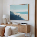 A modern living room with a beige sofa, wooden sideboard, white lamp, stacked books, and the Shifting Tides Canvas Art - Horizontal on a white wall creates a serene atmosphere.