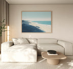 A modern living room features a light beige sectional sofa, a low wooden coffee table, and the Shifting Tides Canvas Art - Horizontal depicting a coastal landscape above the sofa. Natural light brightens the minimalist space.