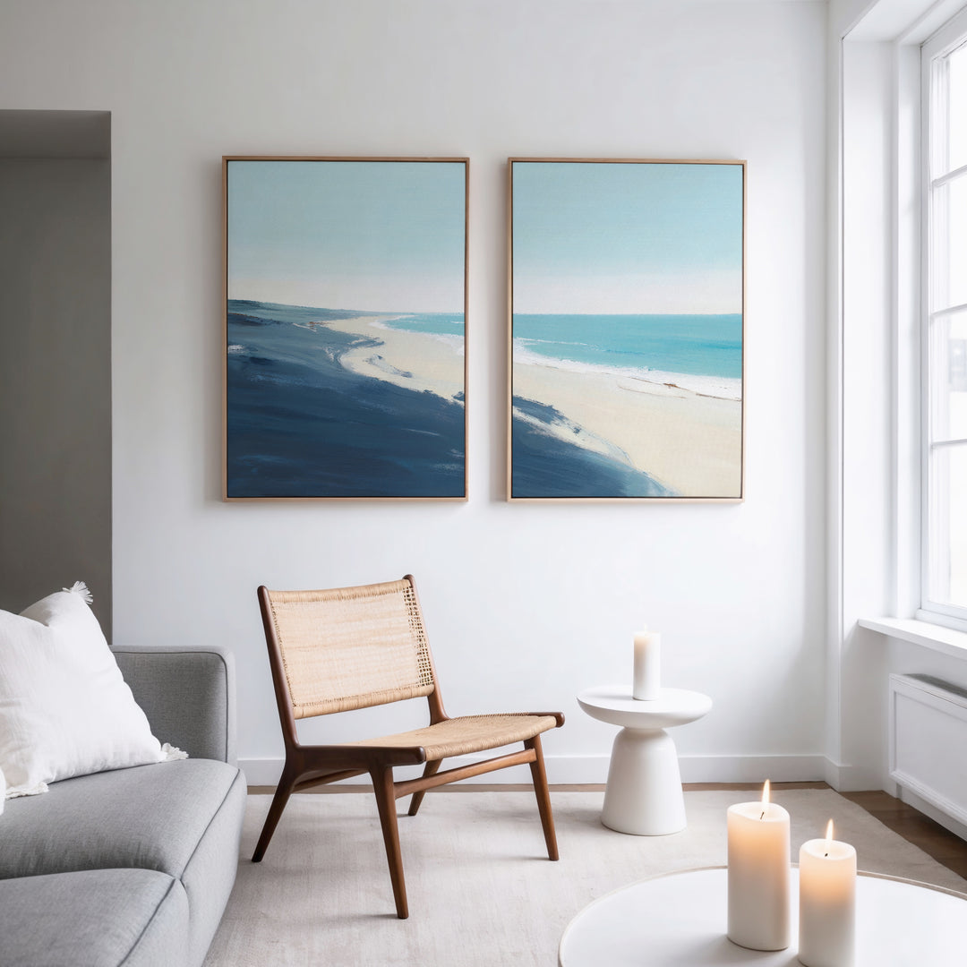 A modern living room features a woven chair, small white table, and lit candles. Above a gray sofa and light wood floors hang the “Shifting Tides - Set of 2” seaside wall art pieces on a white wall.