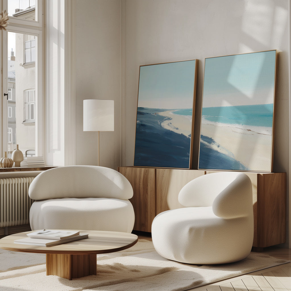 A bright, modern living room features two white rounded armchairs, a low wooden coffee table, and the Shifting Tides - Set of 2 canvas prints leaning above a sideboard. Sunlight through tall windows highlights the serene seaside artwork.