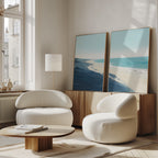 A bright, modern living room features two white rounded armchairs, a low wooden coffee table, and the Shifting Tides - Set of 2 canvas prints leaning above a sideboard. Sunlight through tall windows highlights the serene seaside artwork.