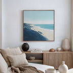 A modern living room featuring a beige sofa, neutral cushions, a wooden sideboard with vases and books, and the Shifting Tides Canvas Art - Square—a large framed coastal landscape print—hanging on the wall.