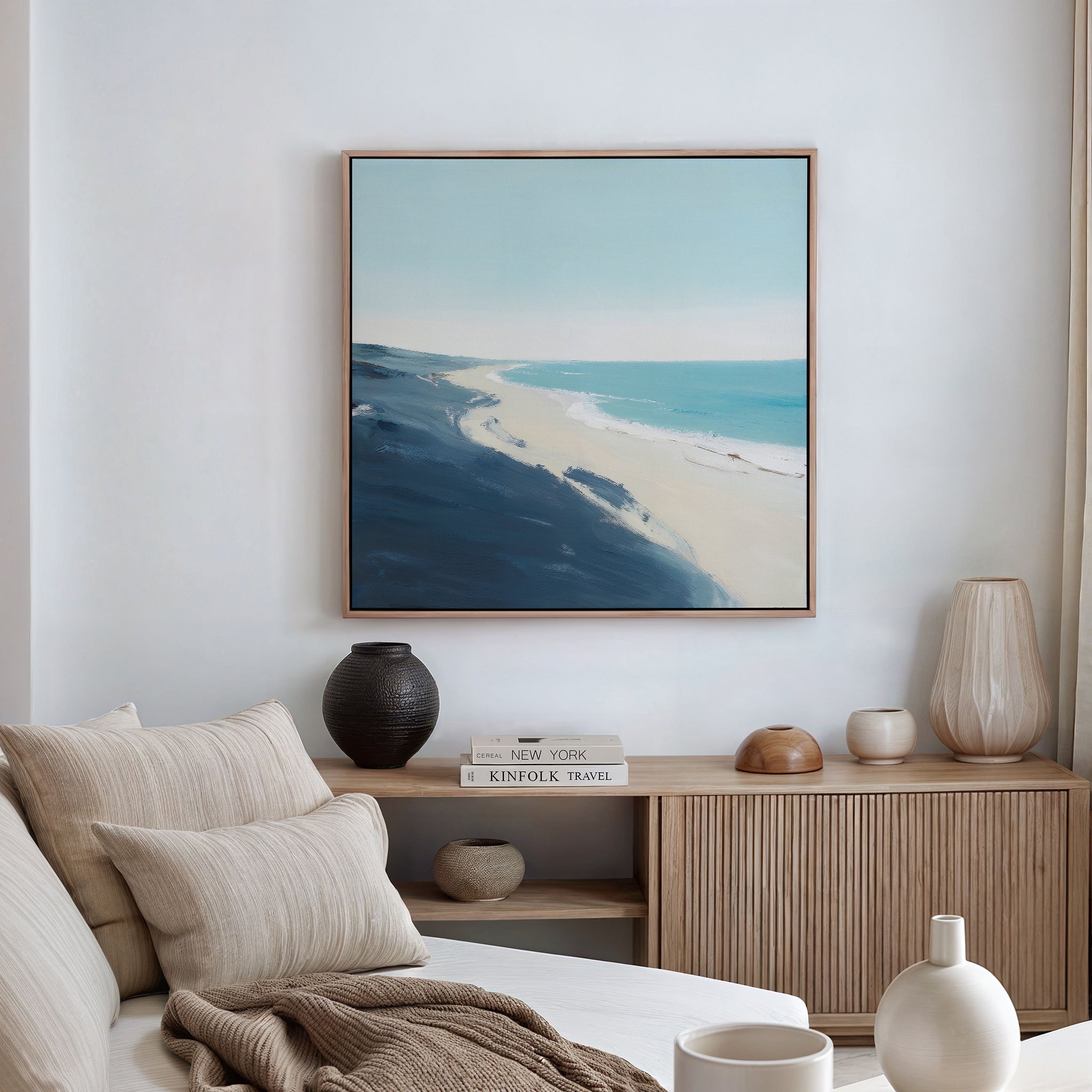 A modern living room featuring a beige sofa, neutral cushions, a wooden sideboard with vases and books, and the Shifting Tides Canvas Art - Square—a large framed coastal landscape print—hanging on the wall.
