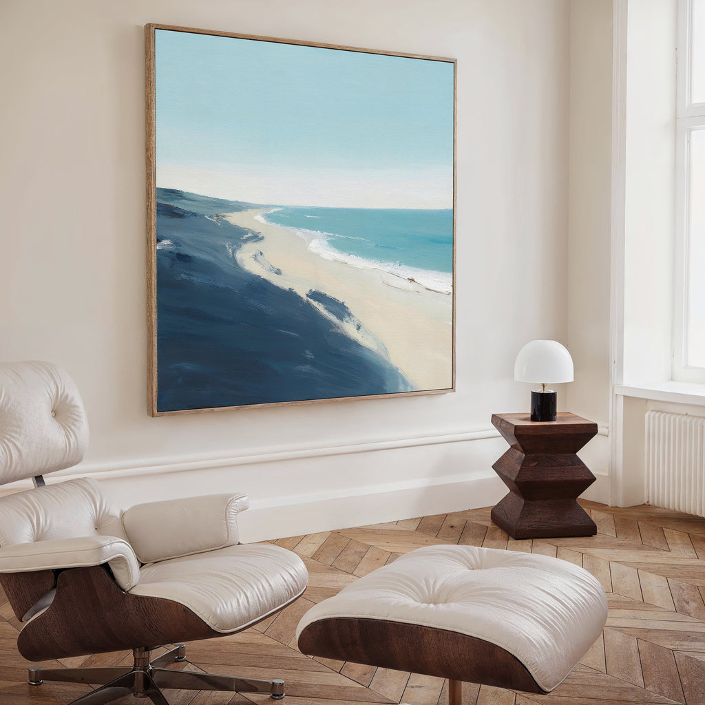 A modern living room featuring a white lounge chair and ottoman, a wooden side table with a small white lamp, and the Shifting Tides Canvas Art - Square depicting a beach scene on the cream-colored wall.