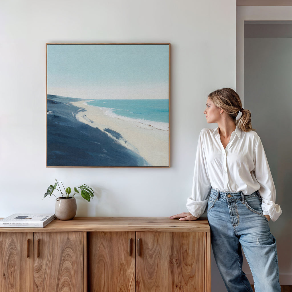 A woman in a white blouse and jeans leans against a wooden cabinet, admiring the Shifting Tides Canvas Art - Square above her. A small potted plant and books sit on the cabinet, giving this Nordic Park-inspired space a cozy touch.