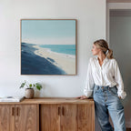A woman in a white blouse and jeans leans against a wooden cabinet, admiring the Shifting Tides Canvas Art - Square above her. A small potted plant and books sit on the cabinet, giving this Nordic Park-inspired space a cozy touch.