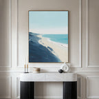 A minimalist room with white paneled walls features a marble console table accented by modern decor and the Shifting Tides Canvas Art - Vertical, a large coastal beach print with blue sky and ocean, hanging above it.