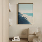 A modern living room corner with a beige sofa, light wood shelves, and the Shifting Tides Canvas Art - Vertical, featuring a beach scene, hanging on a cream wall. A small geometric sculpture is displayed on a lower shelf.