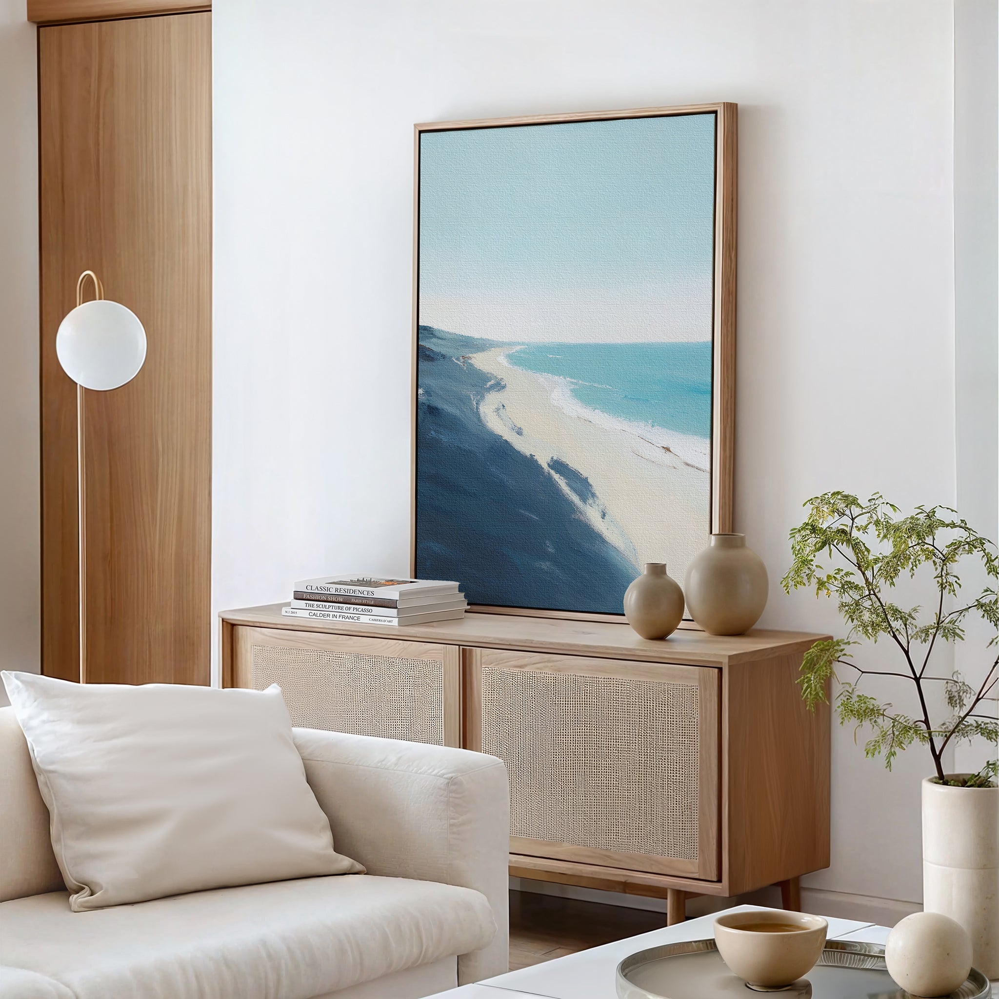 A modern, airy living room features a light wood sideboard, minimalist decor, a potted plant, and the Shifting Tides Canvas Art - Vertical as a large coastal print. In the foreground, a white sofa with pillows adds Nordic Park-inspired relaxation.