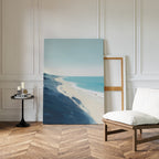 The Shifting Tides Canvas Art - Vertical, featuring a beach with blue waves and sandy shore, leans against a white-paneled wall in a bright room with herringbone floors, a cushioned chair, and a round table with candles and books.