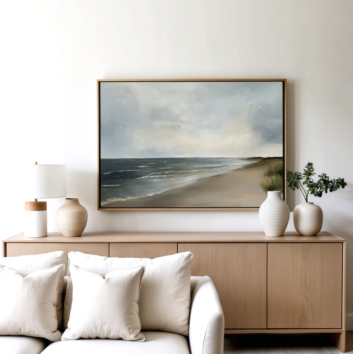 A minimalist living room features a light wood sideboard with white and beige vases, a lamp, and the Shoreline Calm Canvas Art displayed above. A white sofa with neutral pillows completes the serene coastal decor.